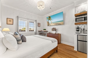 A bedroom with a large bed, a desk, a kitchenette, and a beach-themed painting.