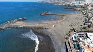A coastal beach with a rocky shoreline, a sandy beach area, and a promenade lined with buildings and palm trees. - Home2Book Disco Duplex in Las Américas Beach (Arona)
