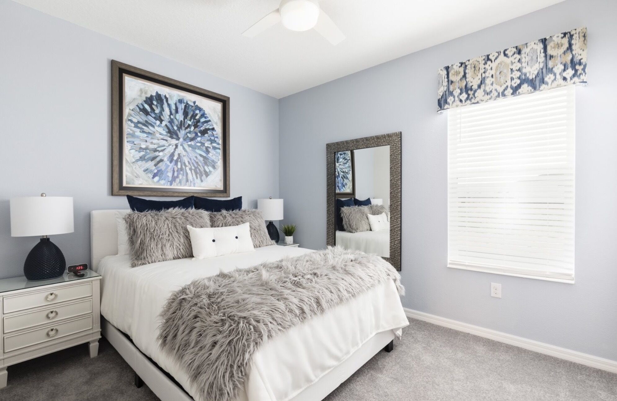 A bedroom with a bed, two bedside tables, a nightstand, a ceiling fan, a large mirror, and a window with blinds.