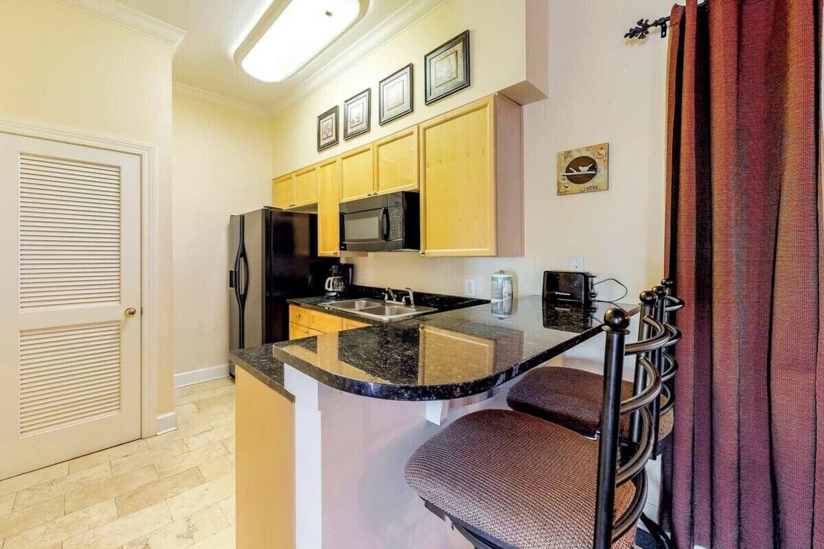 A compact kitchen with a refrigerator, microwave, and sink. There are wooden cabinets and a small island with a sink. A chair is visible near the counter.