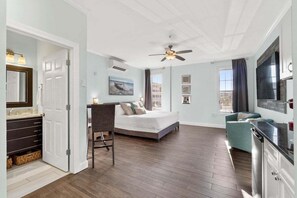 Un dormitorio con cama, escritorio, silla, televisor y baño con espejo y lavamanos.