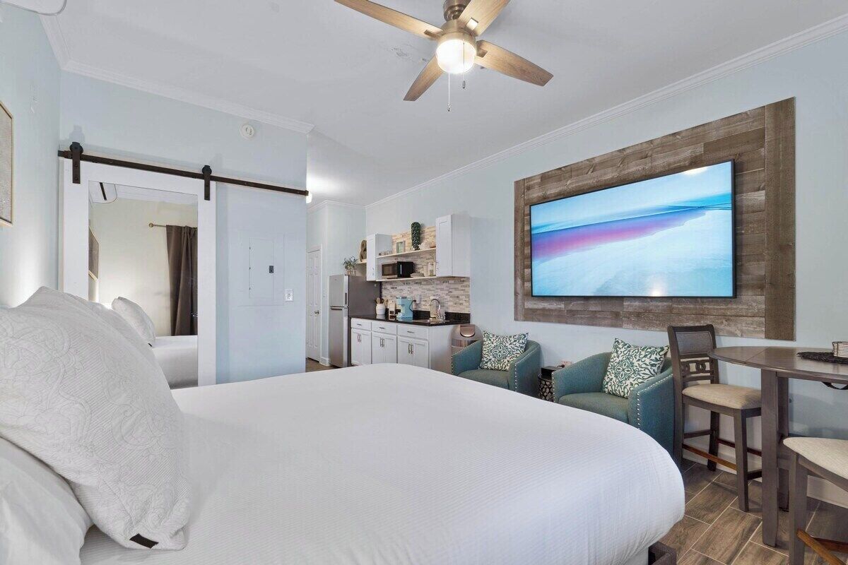 A modern hotel room with a large bed, a dining area, a kitchen, and a TV mounted on the wall.