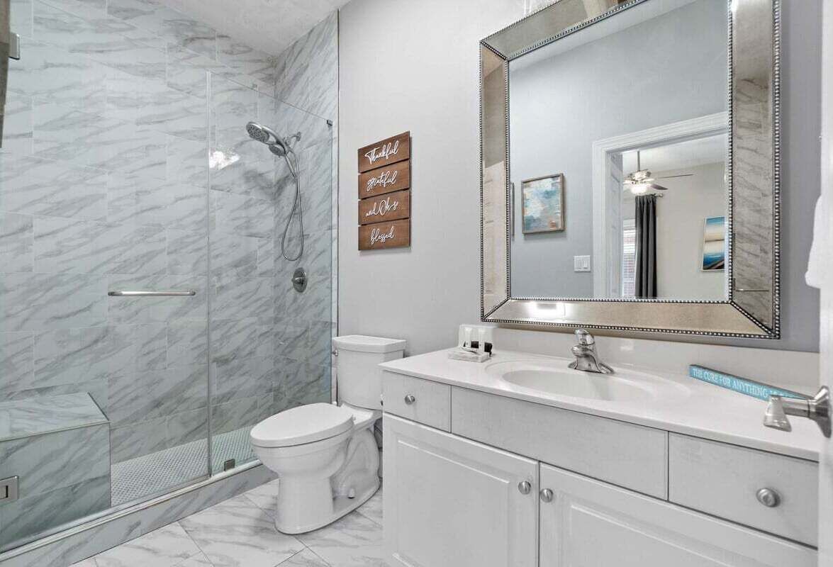 Un baño moderno con cabina de ducha de vidrio, inodoro blanco, tocador con un espejo grande y un cartel decorativo sobre el lavabo.