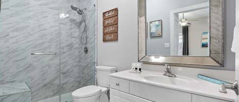 Un baño moderno con cabina de ducha de vidrio, inodoro blanco, tocador con un espejo grande y un cartel decorativo sobre el lavabo.