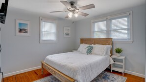 Un dormitorio con cama de madera, ropa de cama blanca, ventilador de techo y dos ventanas con persianas.