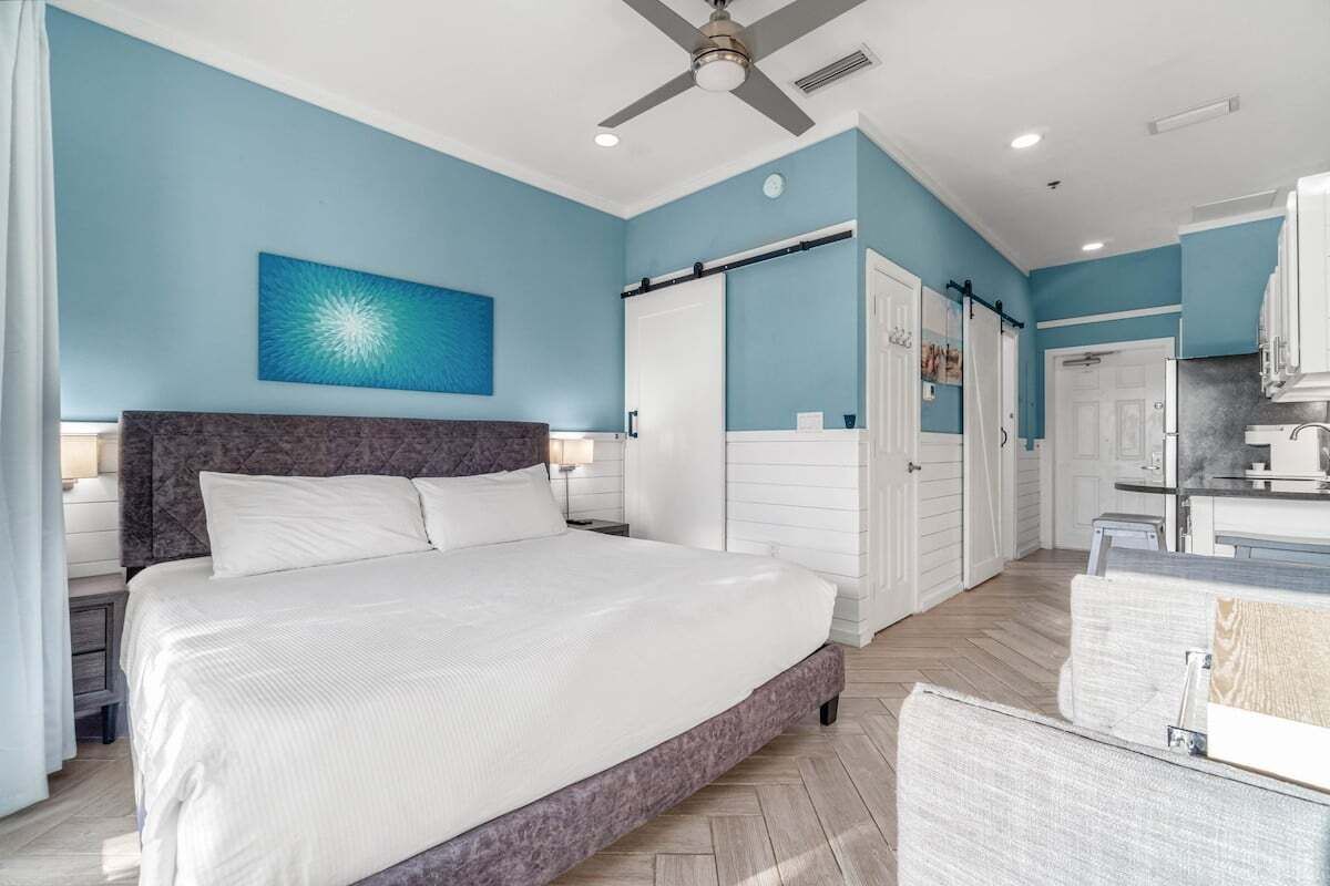 A bedroom with a large bed, a ceiling fan, and a view of the bathroom and kitchen.