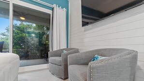A modern room with two grey upholstered chairs, a white bed, and a large sliding glass door leading to a balcony with a view of greenery. - Beach Happy 100-steps to beach-Sleeps 2 (Seacrest)