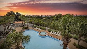 Ein Swimmingpool mit Liegestühlen und Palmen, umgeben von Gebäuden und einem Sonnenuntergangshimmel.
