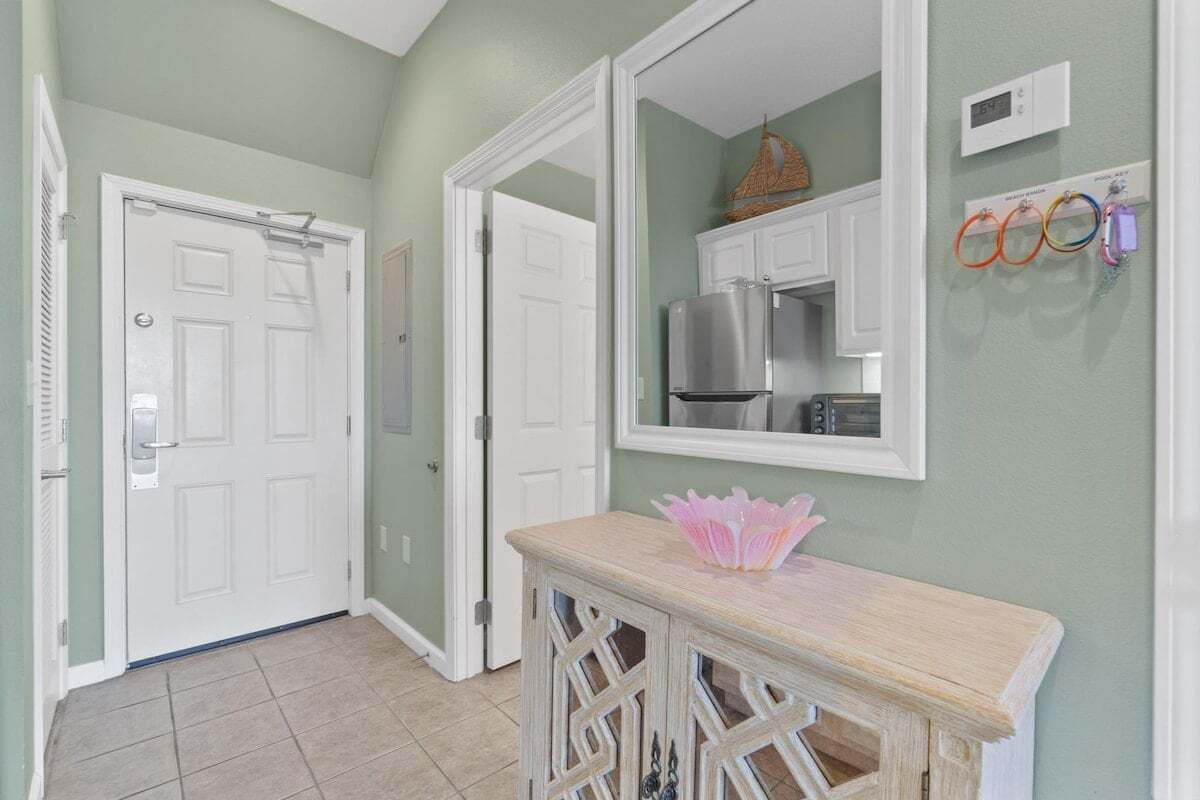 Un pasillo con una puerta blanca, un espejo que refleja una zona de cocina y una pequeña mesa con un adorno.