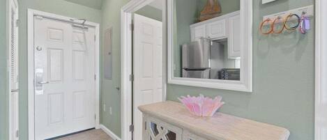 Un pasillo con una puerta blanca, un espejo que refleja una zona de cocina y una pequeña mesa con un adorno.