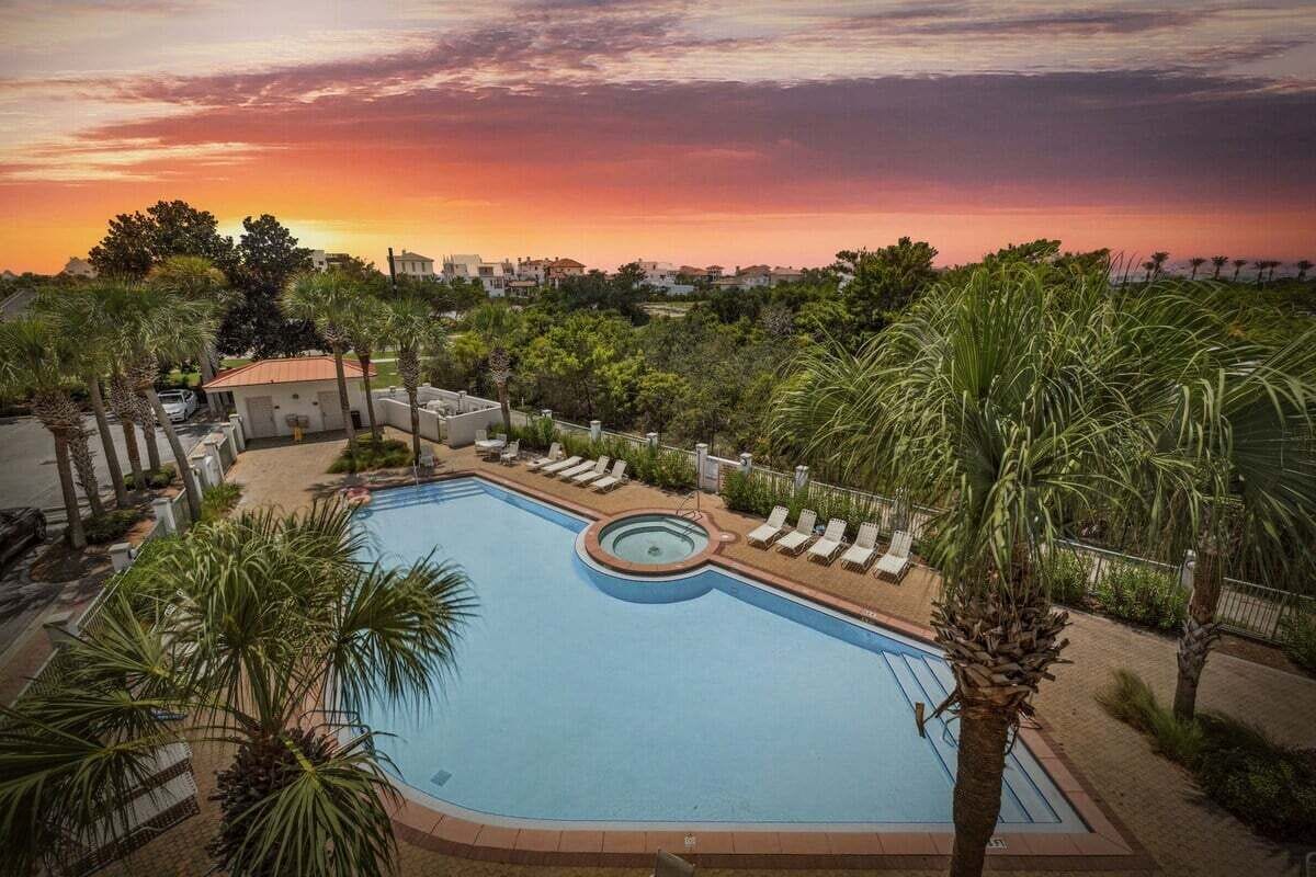 Une piscine entourée de bâtiments, avec des chaises longues et des palmiers, le tout baigné par la lumière d’un coucher de soleil.