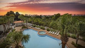 Piscina con sillas de descanso y palmeras, rodeada de edificios y un cielo al atardecer.