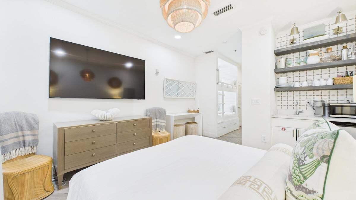A modern bedroom with a large bed, a TV, a dresser, and a kitchen area with shelves.