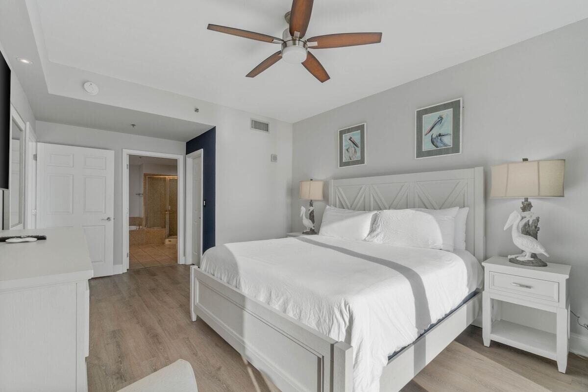 A bedroom with a large bed, a ceiling fan, two bedside tables with lamps, and a door leading to another room.