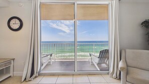 A room with a view of the ocean through a sliding glass door, a clock on the wall, and a chair by the door.