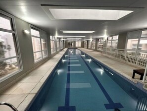 Uma piscina interior comprida com água azul cristalina, rodeada por janelas e soalho em azulejos.