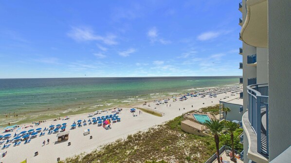 A beach with rows of sun loungers and umbrellas, a pool, and a view of the ocean.