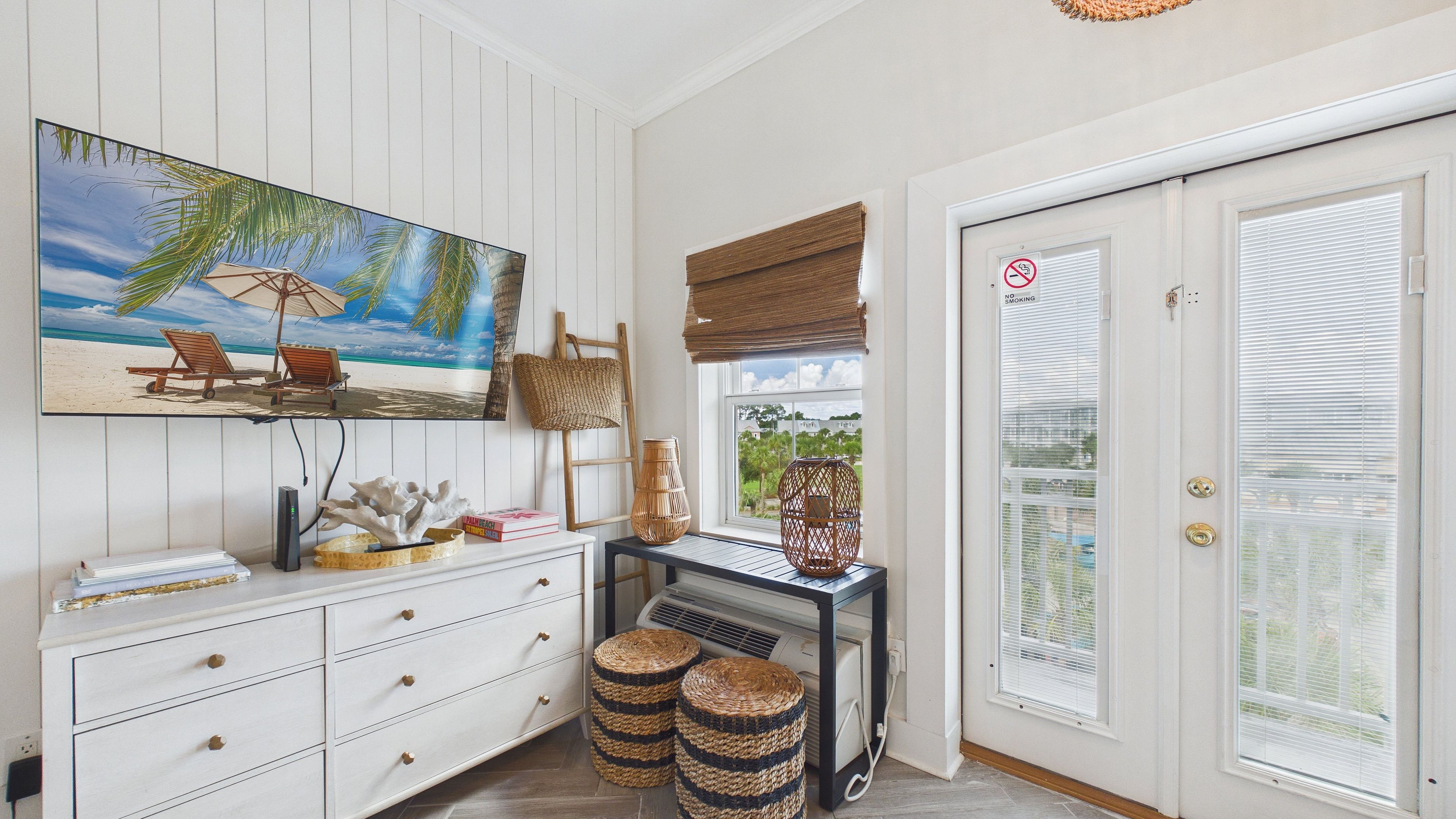 A room with a beach-themed painting, a white dresser, wicker baskets, and a door with a 'No Smoking' sign.