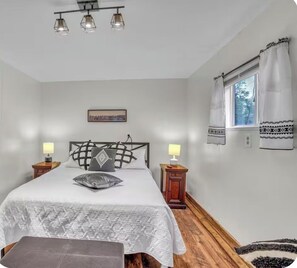 Un dormitorio con cama, lámparas de noche, ventana con cortinas, un cuadro en la pared y suelo de madera.