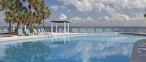 A swimming pool with lounge chairs and palm trees, surrounded by a fence and a clear sky.