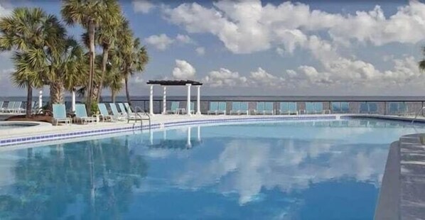 Une piscine entourée de chaises longues et de palmiers, le tout délimité par une clôture, sous un ciel dégagé.