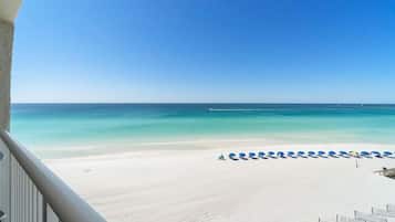 Vista a la playa desde un balcón, con una fila de sombrillas azules y sillas reclinables blancas.