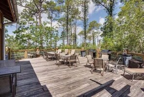An outdoor deck with wooden flooring, metal chairs, and tables, surrounded by trees.