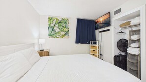 Une chambre d’hôtel comprenant un lit, une table de chevet, un téléviseur fixé au mur, une armoire avec des vêtements suspendus et un ventilateur.