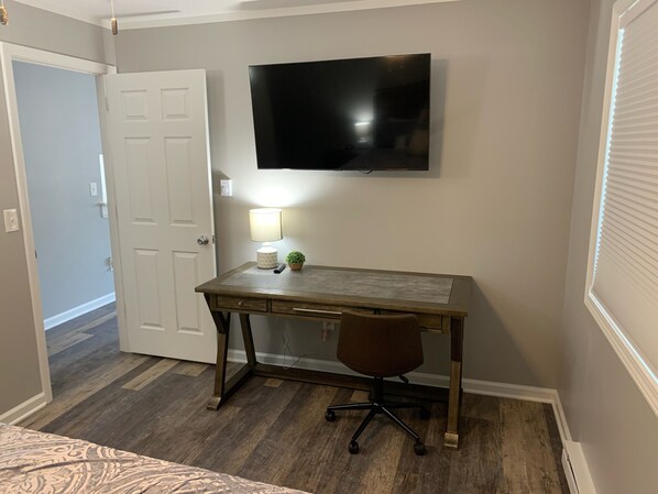 A bedroom with a wooden desk, a chair, a TV mounted on the wall, and a door leading to another room.