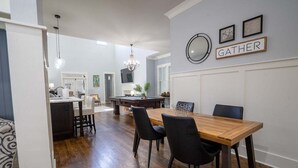 Un comedor con una mesa de madera y sillas negras, una araña y un letrero que dice 'GATHER' en la pared.