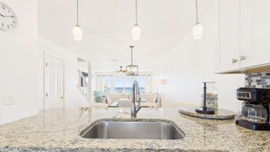A kitchen with granite countertops, a stainless steel sink, pendant lighting, and a view to the living area.