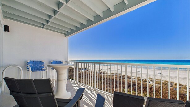 A balcony with a view of the beach, white railing, and a table with chairs.