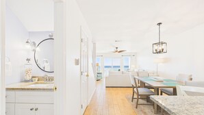 A bright, spacious room with a wooden floor, a dining area with a table and chairs, a large window with a view of the sea, and a ceiling fan.