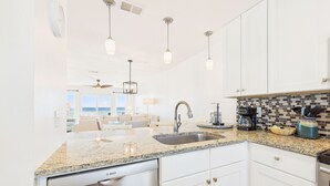 A modern kitchen with white cabinets, granite countertops, and stainless steel appliances. There is a dining area with a view of the ocean.