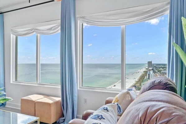 A room with a large window offering a view of the beach and ocean. There are two cushioned seating areas and a small table. - Sea Esta 1900–Gulf Views 2BR–Sleeps 6 (Panama City Beach)