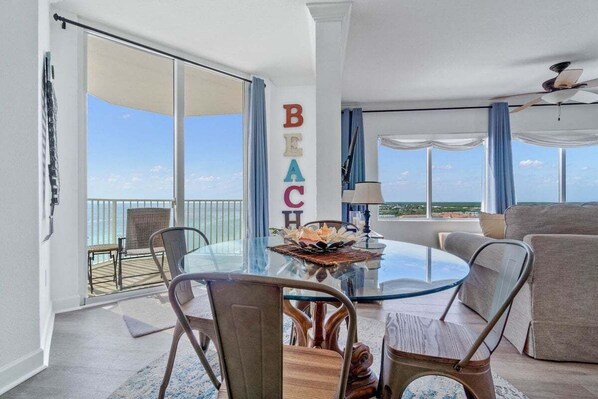 A spacious living room with a glass table, comfortable seating, a balcony with a view, and a decorative word wall spelling "BEACH".