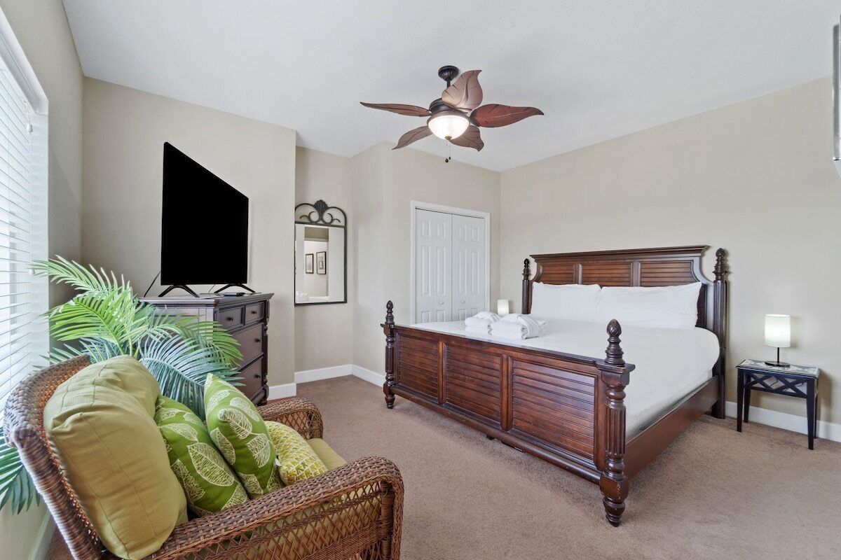 A bedroom with a wooden bed, a ceiling fan, a dresser, a television, and a wicker chair.