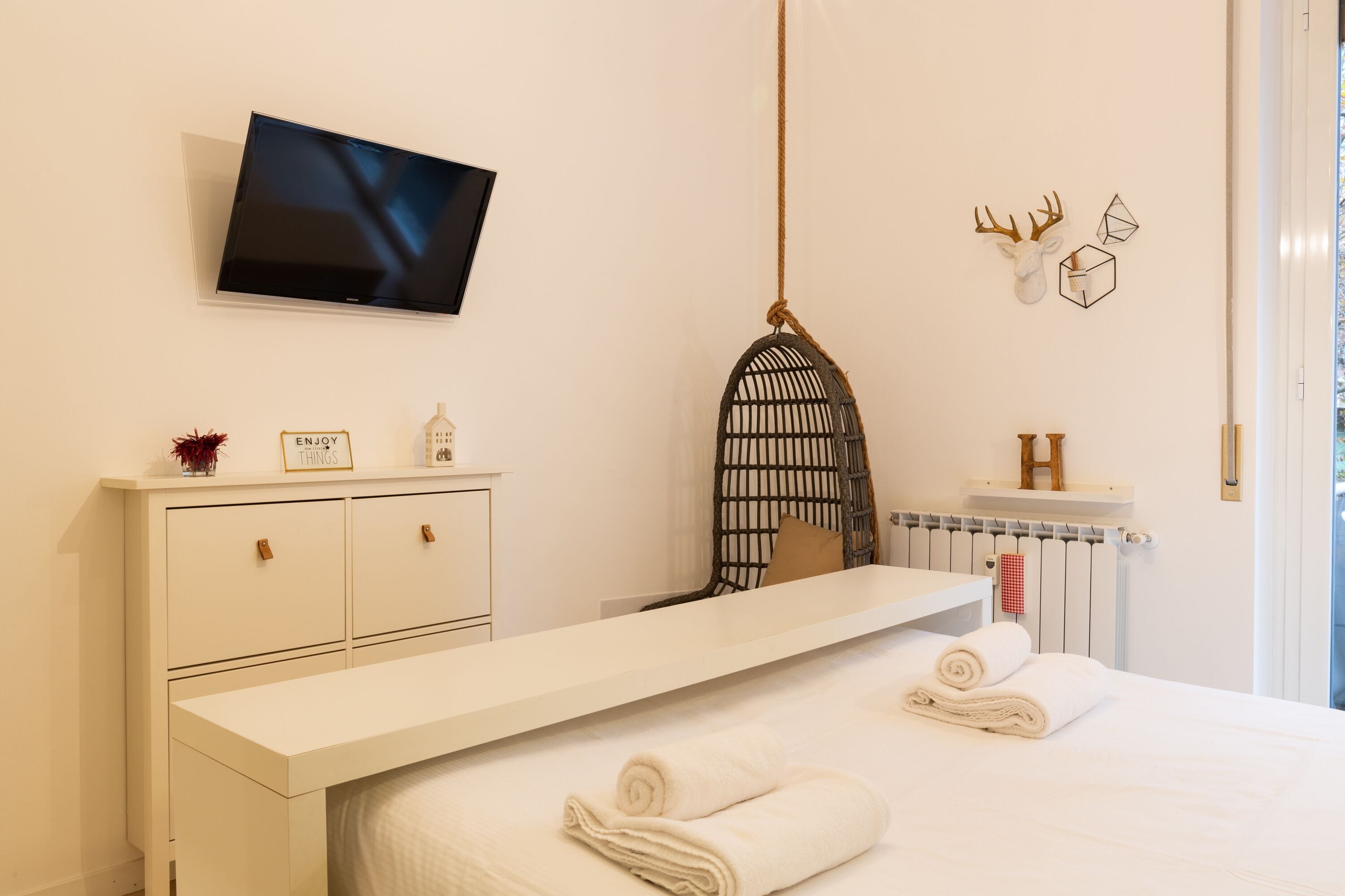 A modern hotel room with a large bed, a flat-screen TV mounted on the wall, a white dresser, and a unique wall decor piece.