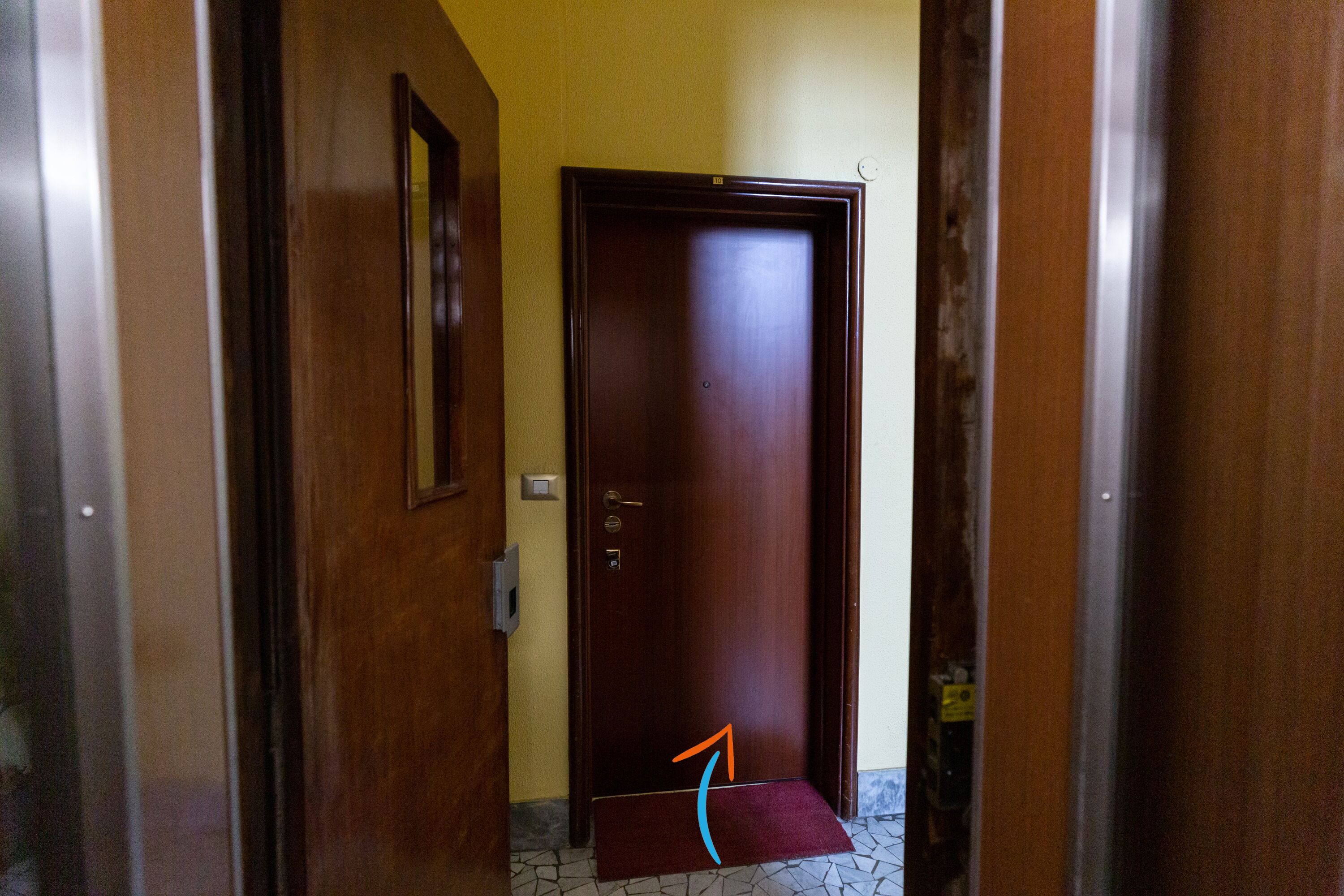 A wooden door with a doormat, a light switch, and a wall-mounted light fixture.
