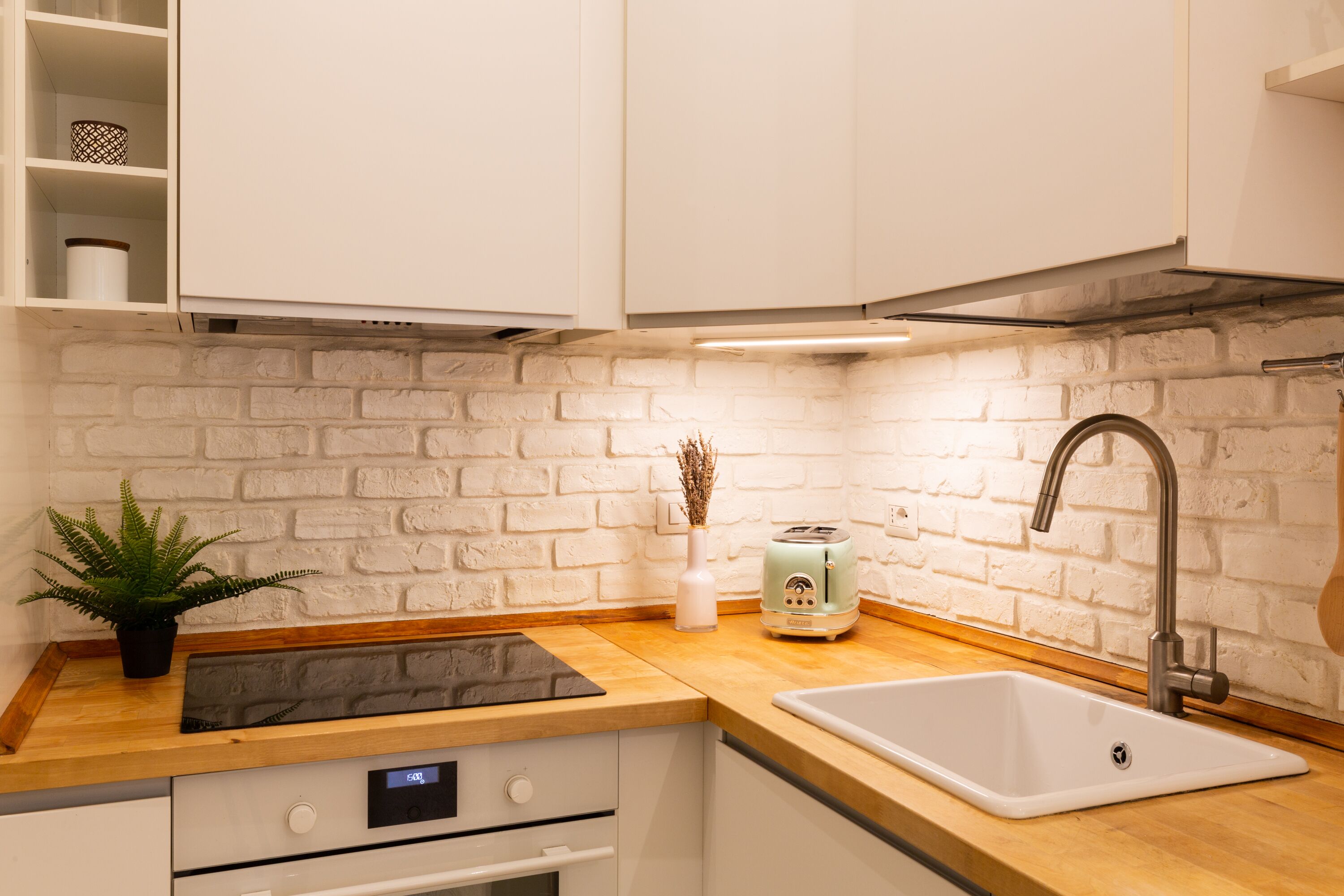 Uma cozinha com armários brancos, backsplash de parede de tijolos, pia, torradeira e um vaso com planta.