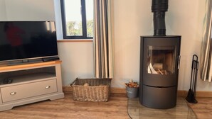 A modern living room with a TV on a wooden stand, a black wood-burning stove, a wicker basket, and a glass table. - Luxury Secluded Barn With Private Hot Tub (Denbigh)