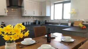A modern kitchen with white cabinets, a wooden dining table set with white plates and bowls, a bottle of wine, and a vase of yellow flowers. - Luxury Secluded Barn With Private Hot Tub (Denbigh)
