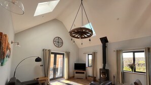 A spacious living room with a large black leather sofa, a wooden ceiling, a black wood-burning stove, and a large window with curtains. - Luxury Secluded Barn With Private Hot Tub (Denbigh)