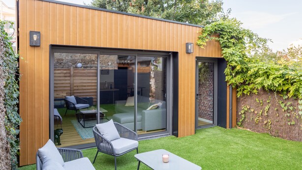 En moderne udendørs terrasse med træterrasse, en glas skyde dør ind til et møbleret interiør, og et lille bord med et stearinlys.
