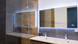 Et moderne badeværelse med et brusebad i glas, et stort spejl og en trækommode med to vaske.