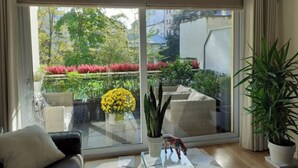 A room with a large sliding glass door leading to a garden with various plants and flowers. There is a sofa and a small table with a decorative item on it. - Very nice apartment downtown, Parc Olympe de Gouges ( with garage) (Annemasse)