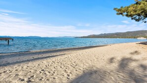 Une plage avec une jetée en bois s’avançant dans des eaux bleues et calmes, une rive sablonneuse et des montagnes lointaines.