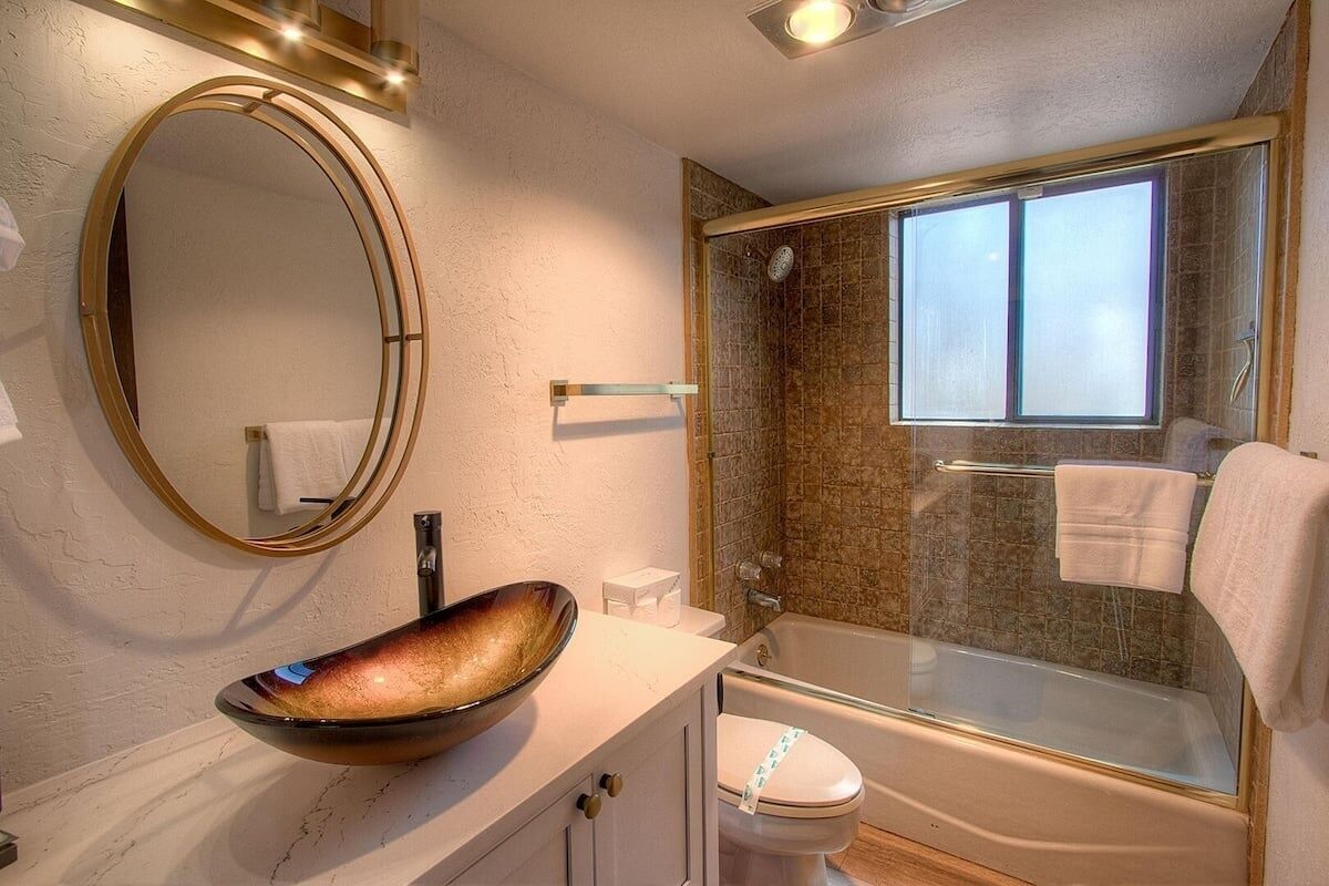 A bathroom with a round mirror, a vessel sink, a toilet, and a glass-enclosed shower area.