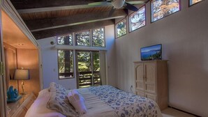 Un dormitorio con una cama, un televisor sobre un mueble y una ventana con vistas a árboles.