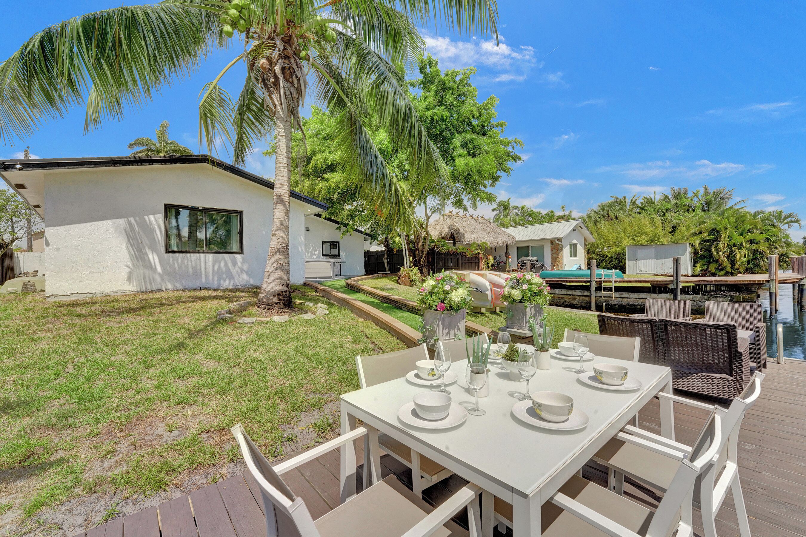 A dining area with a white table set for four, surrounded by wicker chairs, with a pool and a house in the background.